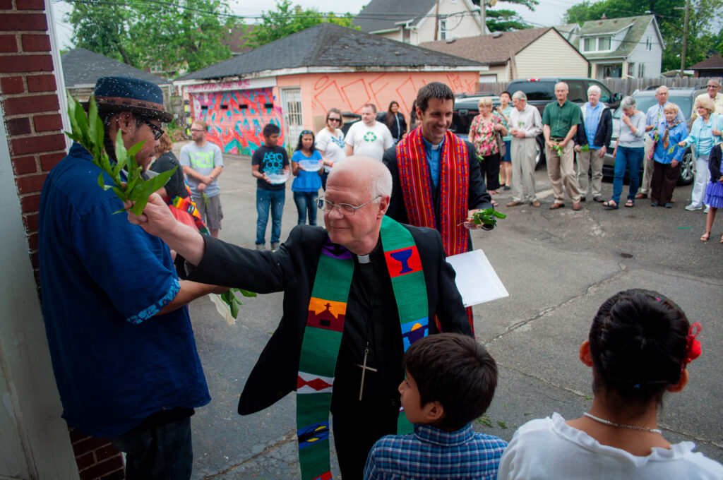 Pastor Jack Eggleston and Pastor John bless Grace in Action’s new space with holy water in June 2025, joined by community members gathered outside.