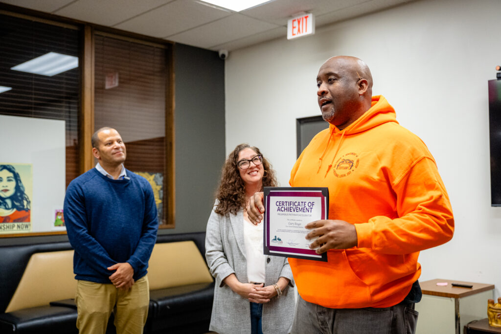 Gary Ringer holds a certificate while speaking at a ProsperUs Construction Accelerator graduation event. Founder of Detroit Future Ops, Tanya Saldivar-Ali, ProsperUs Detroit CEO, Paul Jones, stand beside him.