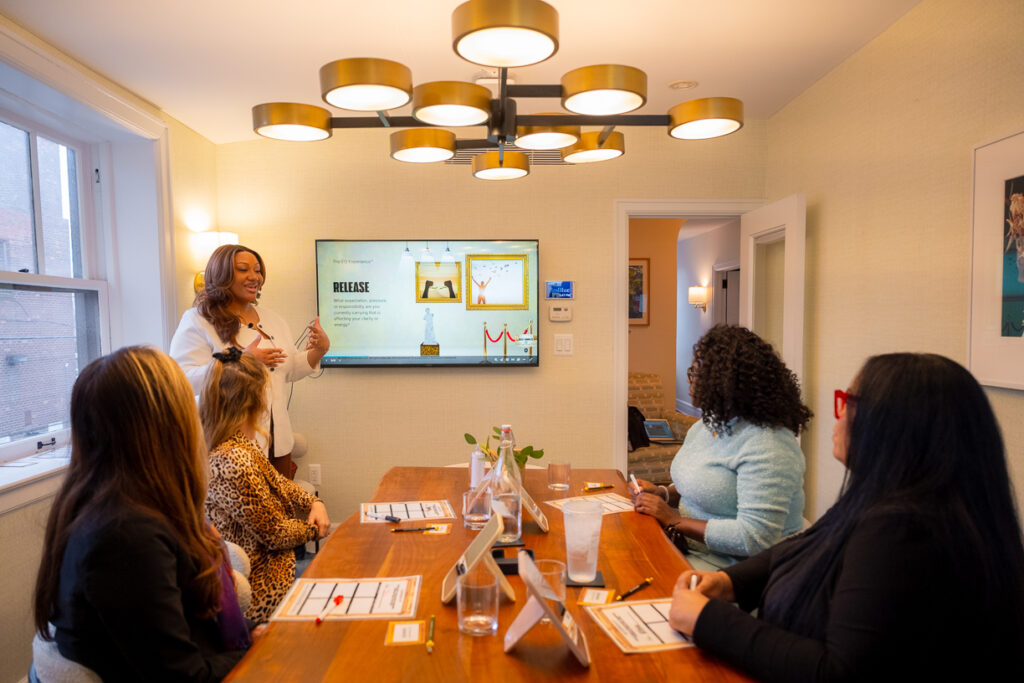 Tiara B.A. leads a small group workshop, presenting on emotional release and wellness concepts to a group of women seated around a table.