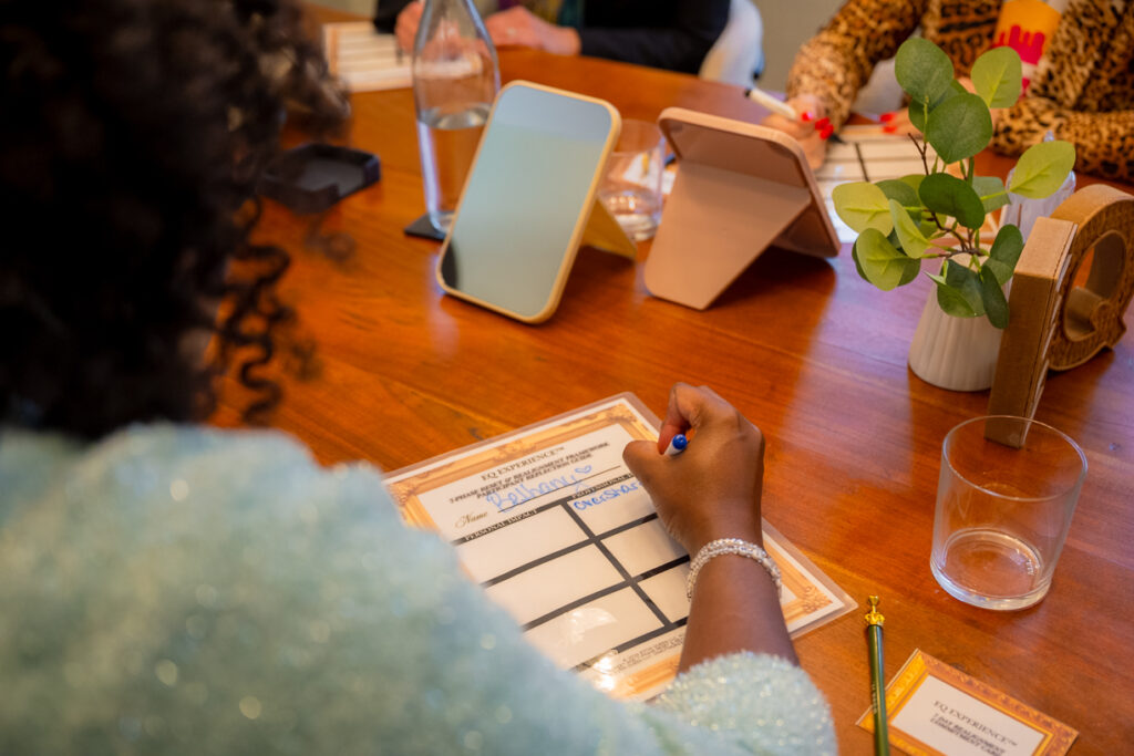 Participant writes on an EQ Experience worksheet during an Esthi Queen session, with mirrors and materials arranged on a wooden table.
