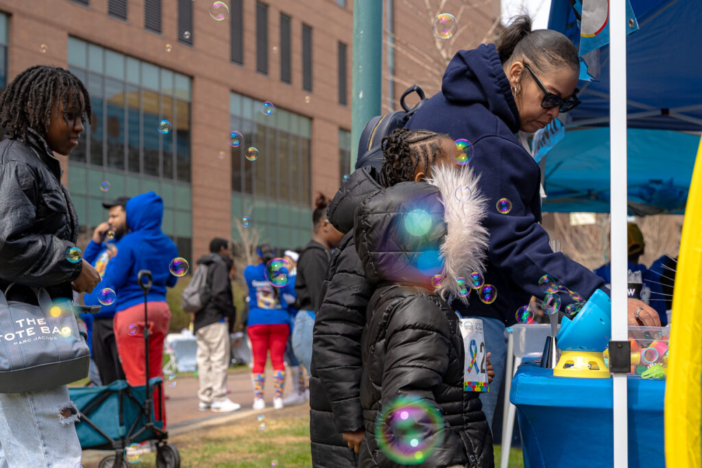 Child and adult interact at event booth as bubbles float, surrounded by families at Autism Awareness Walk.